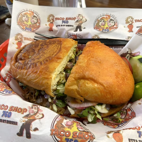 A large Mexican torta sandwich, cut in half, rests in a red basket lined with Taco Shop 760 paper.