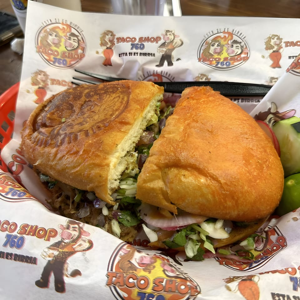 A large Mexican torta sandwich, cut in half, rests in a red basket lined with Taco Shop 760 branded paper.